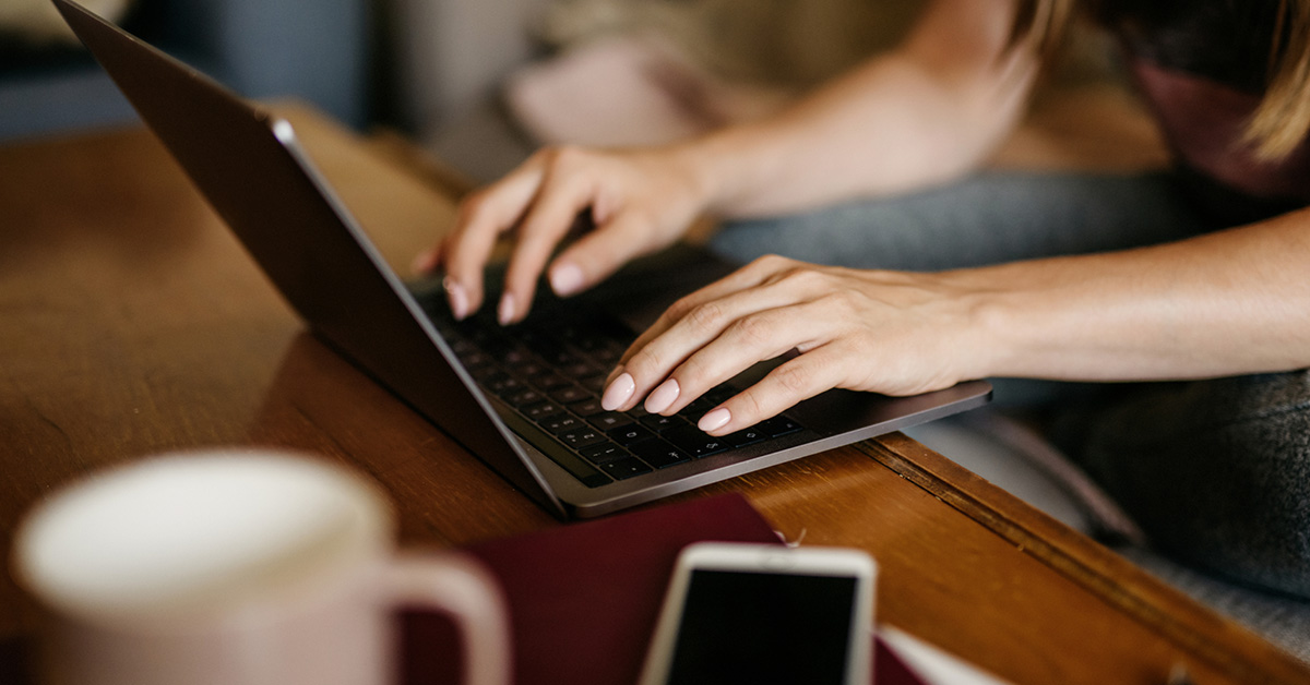 Hands typing on a laptop with a smartphone and coffee mug on the table.