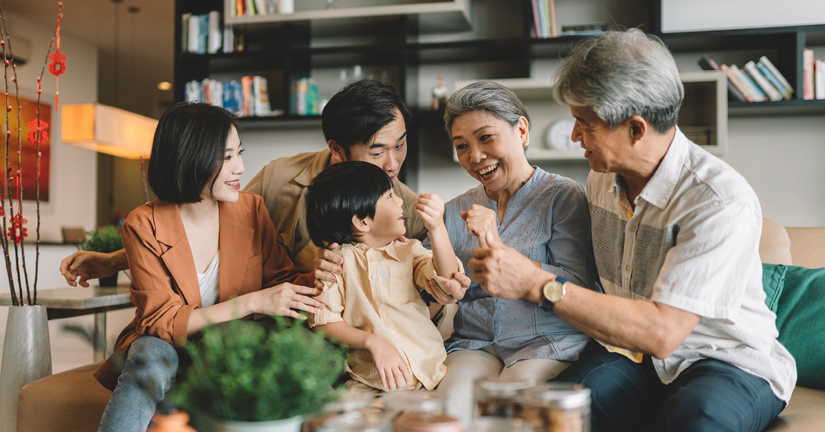 Four middle-aged people in office casual attire sit around a table in a pleasant, modern office break room flooded with natural light enjoying a business breakfast. A woman holds court as the other three listen attentively, two of them with tablets open in front of them.