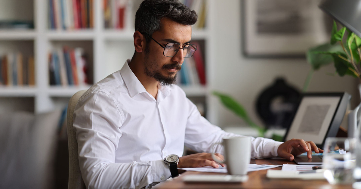 A young man with a beard and glasses sits at a desk in his home office poring over a page of financial information.