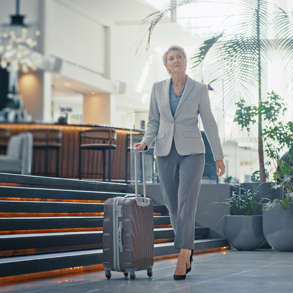 A person in a grey suit is walking through a modern, well-lit indoor space with high ceilings and large windows. The person is holding the handle of a rolling suitcase. In the background, there are bar stools and tables, as well as some plants and decorative lighting fixtures. The overall atmosphere appears to be that of an upscale hotel lobby or airport lounge.